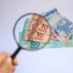 Mão segurando uma lupa em primeiro plano, ampliando cédulas de real espalhadas sobre uma superfície clara. Dentro da lente aparecem com destaque notas de R$10 e R$100, enquanto outras cédulas permanecem desfocadas ao redor. A imagem remete à análise, conferência ou avaliação de valores financeiros. Logotipo da empresa no canto inferior esquerdo.